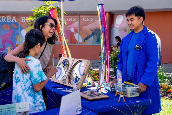 Staff in a blue lab coat interacting with a family at an outdoor science festival.