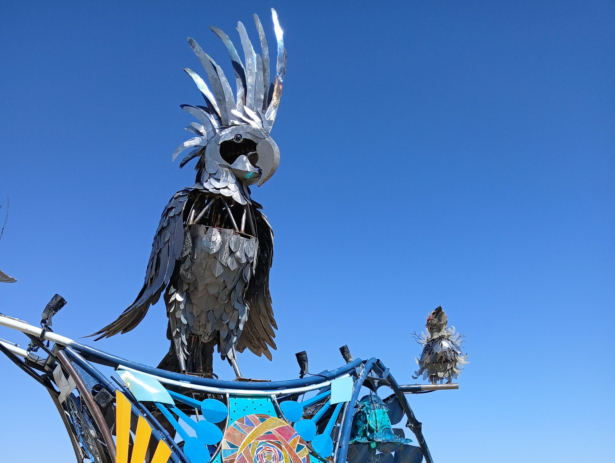 Large metallic bird sculptures with intricate feather details are displayed against a clear blue sky, perched on a colorful structure decorated with mosaic patterns and geometric shapes.