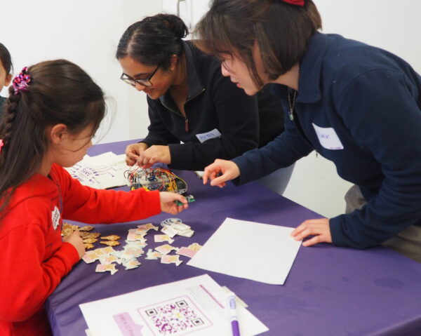 A group of people engaging in a crafting activity at a table covered with a purple cloth. A child is sorting colorful puzzle pieces, and two adults are facilitating the activity. A QR code and worksheets are visible on the table.