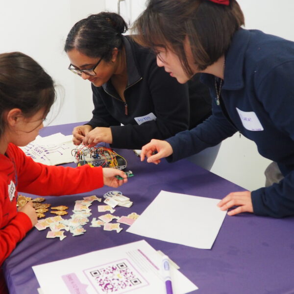 A group of people engaging in a crafting activity at a table covered with a purple cloth. A child is sorting colorful puzzle pieces, and two adults are facilitating the activity. A QR code and worksheets are visible on the table.