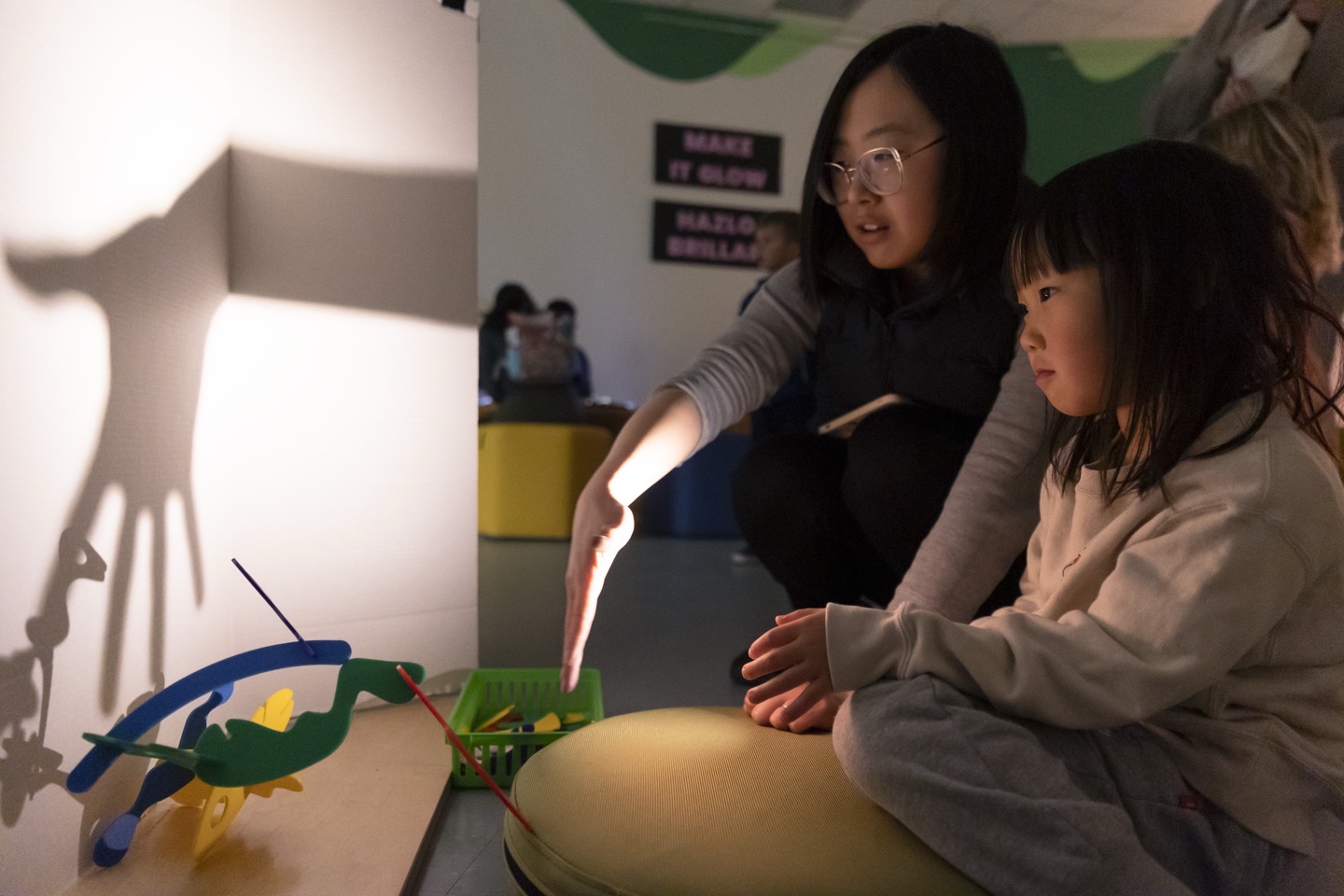 Adult and child creating shadow puppets on a light screen using colorful cutout figures during the Glowtastic exhibit