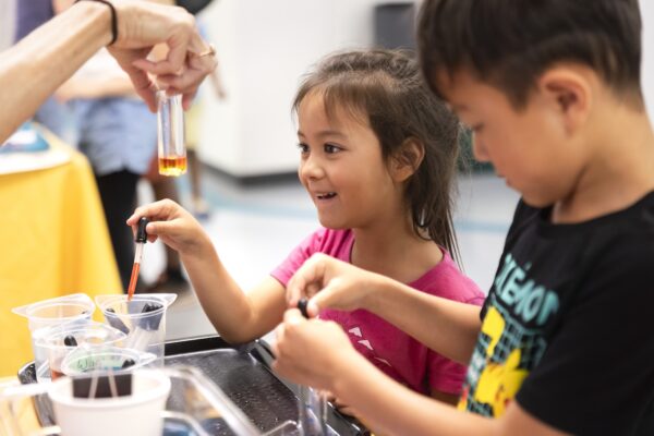 Children engaged in a science activity, using droppers to mix liquids, with an adult holding a test tube nearby.