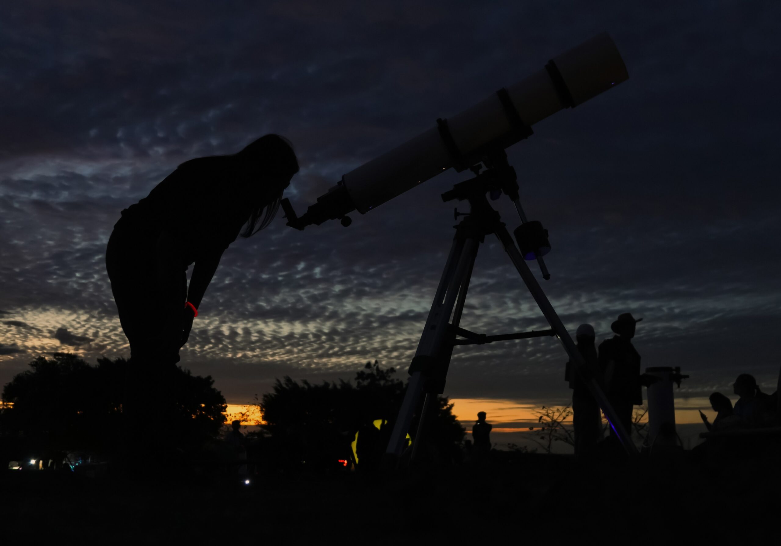 A person is silhouetted against a sunset, looking through a telescope. The sky is filled with dark clouds, and other people and trees are faintly visible in the background.