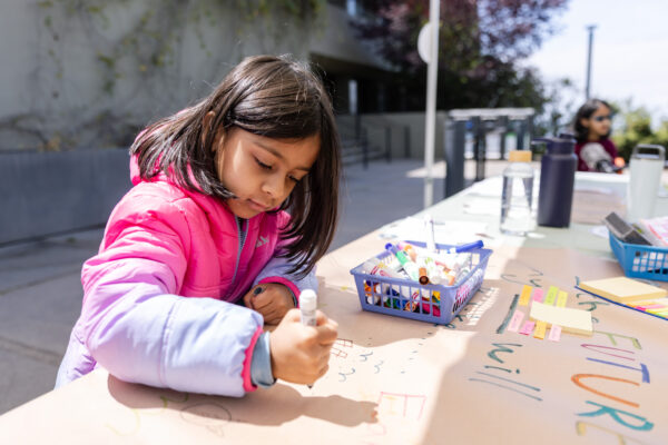 A child wearing a pink jacket is drawing on a large sheet of paper outdoors. The paper is filled with colorful markers and illustrations.