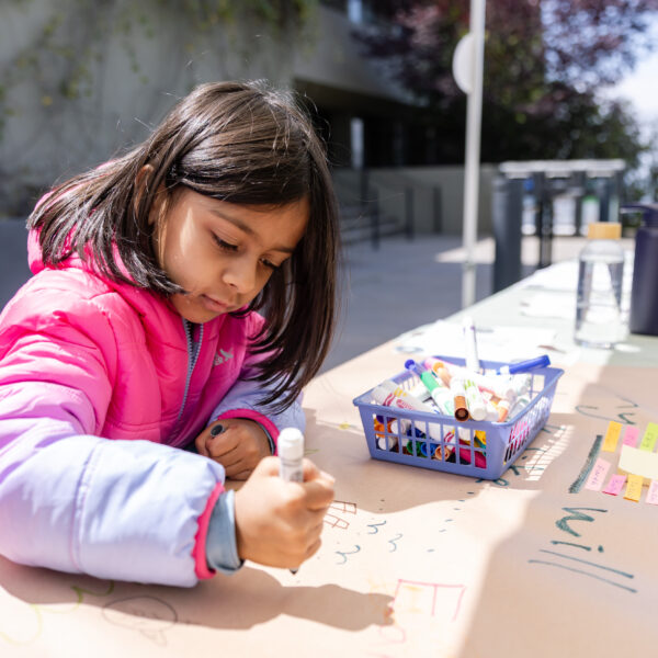 A child wearing a pink jacket is drawing on a large sheet of paper outdoors. The paper is filled with colorful markers and illustrations.
