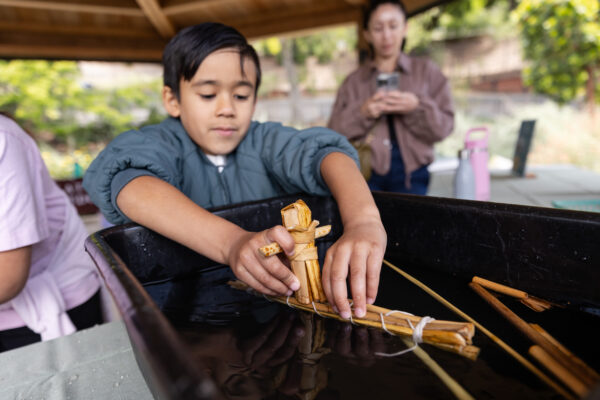 A child carefully places a small tule boat into a black basin filled with water, while another person watches in the background.