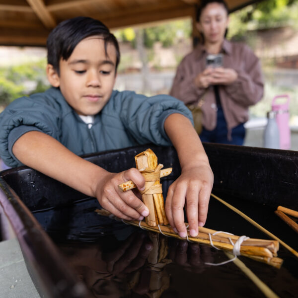 A child carefully places a small tule boat into a black basin filled with water, while another person watches in the background.