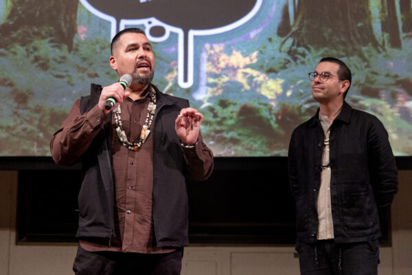 Vincent Medina speaking into a microphone with Louis Trevino listening attentively, with a forest-themed backdrop behind them.