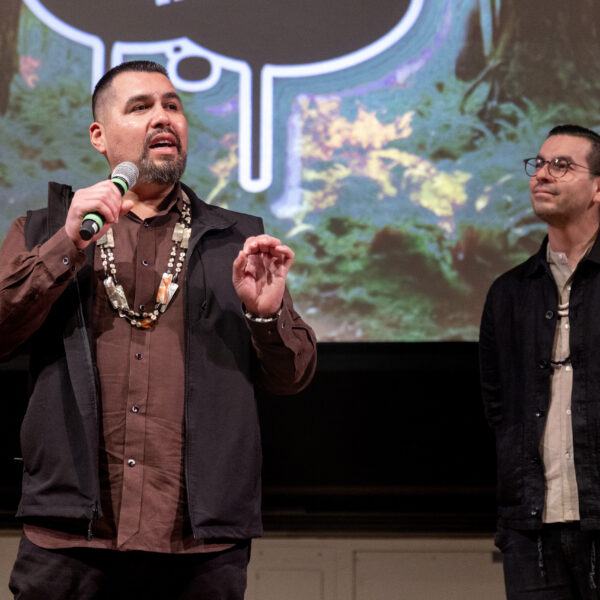 Vincent Medina speaking into a microphone with Louis Trevino listening attentively, with a forest-themed backdrop behind them.