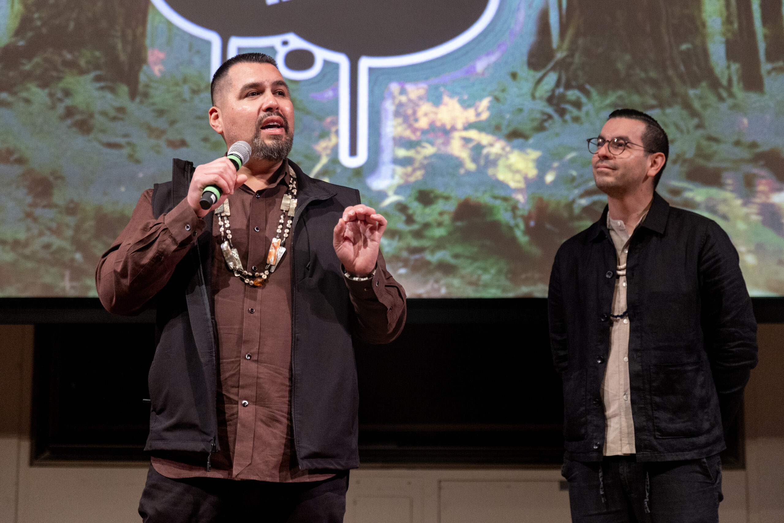 Vincent Medina speaking into a microphone with Louis Trevino listening attentively, with a forest-themed backdrop behind them.