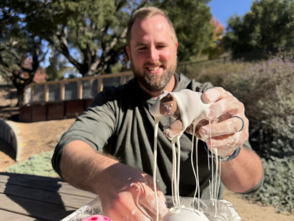 A person outdoors holds a bear toy dripping with oobleck, smiling as gooey strands fall from their hands.